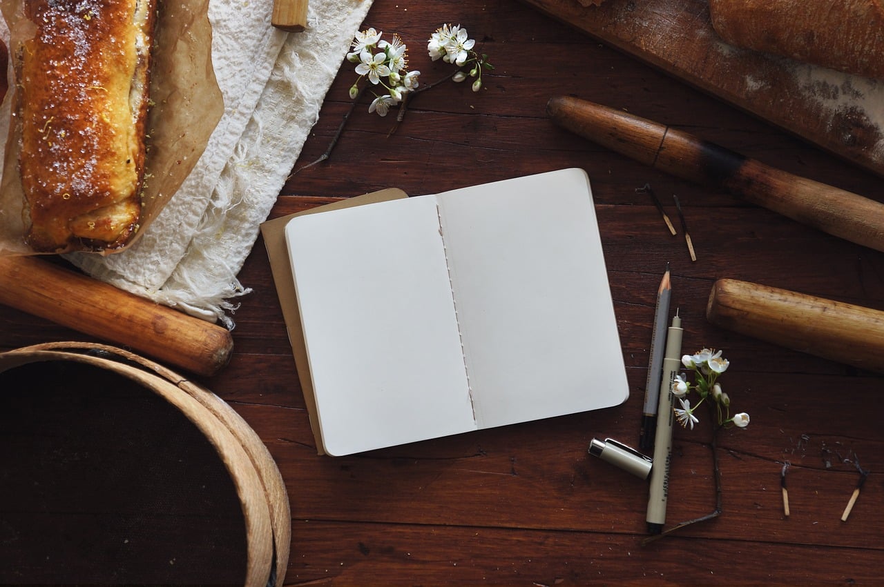 white notebook with pens, flowers and bread around it